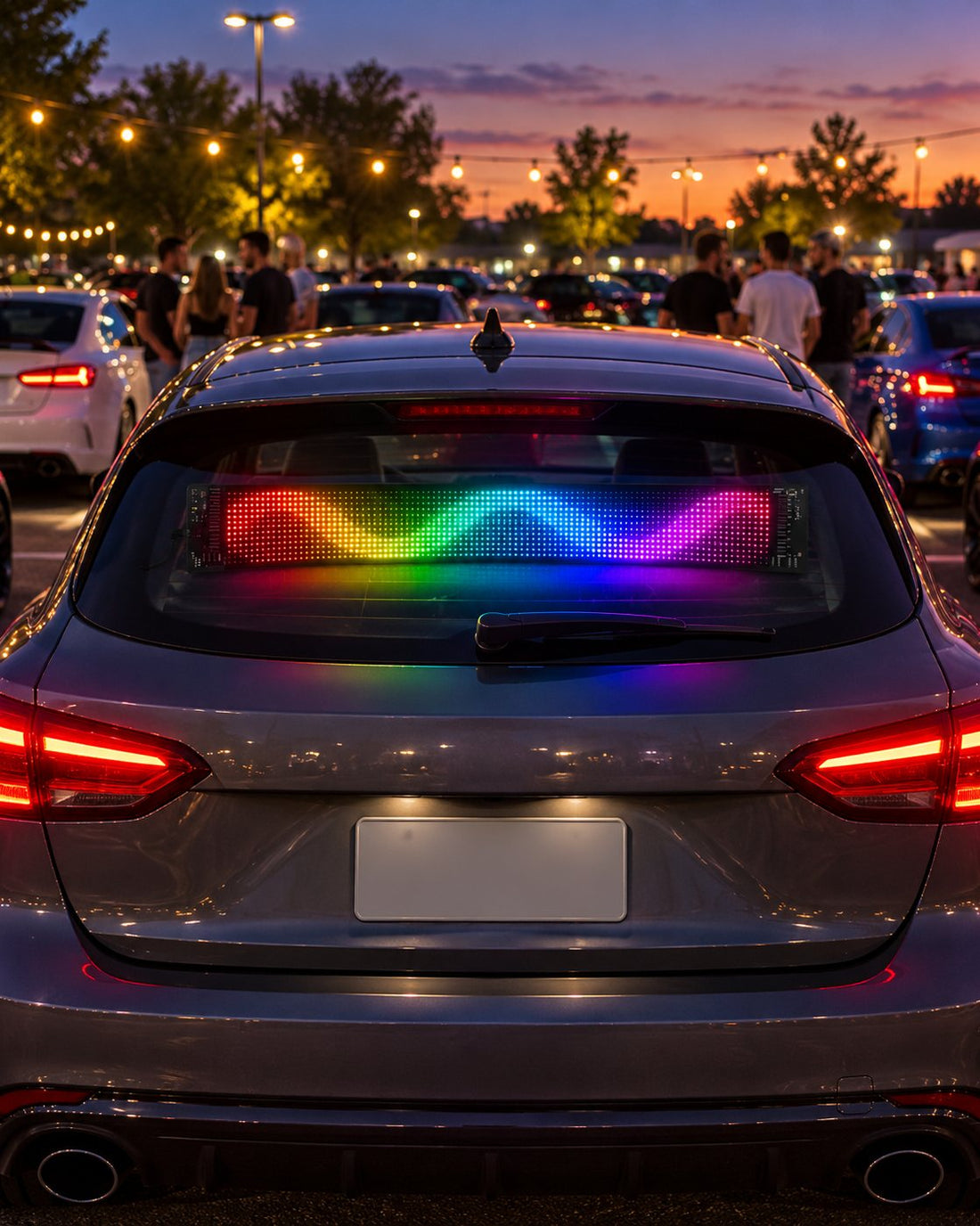 The LED panel in the rear window of a parked car at an evening car meet, showing a rainbow wave